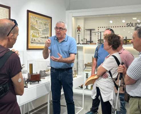 El director del museo explica una pieza a un grupo de visitantes, una de ellas con bastón blanco, junto a una lámina de pesas y medidas del sistema métrico decimal.