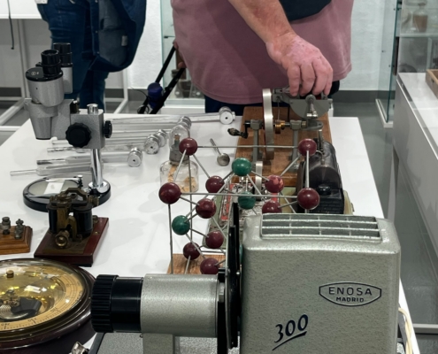 Primer plano de la mano de un visitante sobre una pequeña máquina de vapor, junto a una maqueta de estructura molecular, diapasones, un barómetro y un proyector.