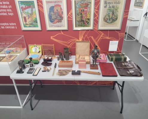 Mesa con objetos escolares preparados para tocar: ábaco, cartera de cuero, plumieres, sacapuntas de manivela, calculadora, regla de cálculo, sellos de madera y un mapa en relieve de España.
