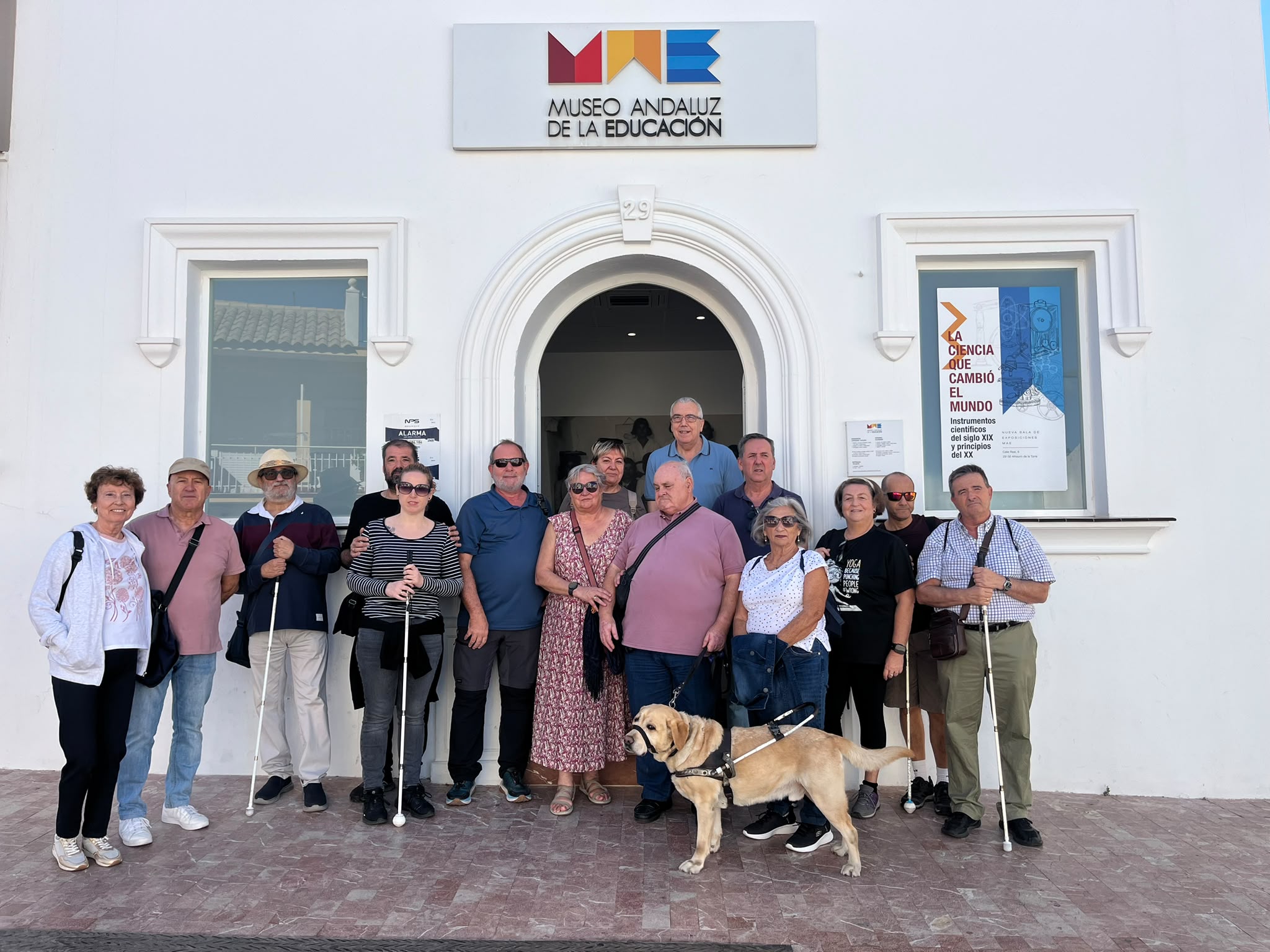 Grupo de visitantes de la ONCE de Málaga, varios con bastón blanco y uno con su perro guía, posando ante la fachada blanca del Museo Andaluz de la Educación junto al director del museo.